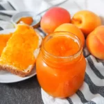 A view of a apricot jam in a glass jar with some jam on the toast and some apricots on the side