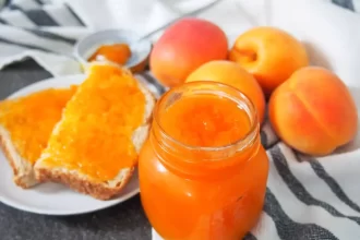 A view of a apricot jam in a glass jar with some jam on the toast and some apricots on the side