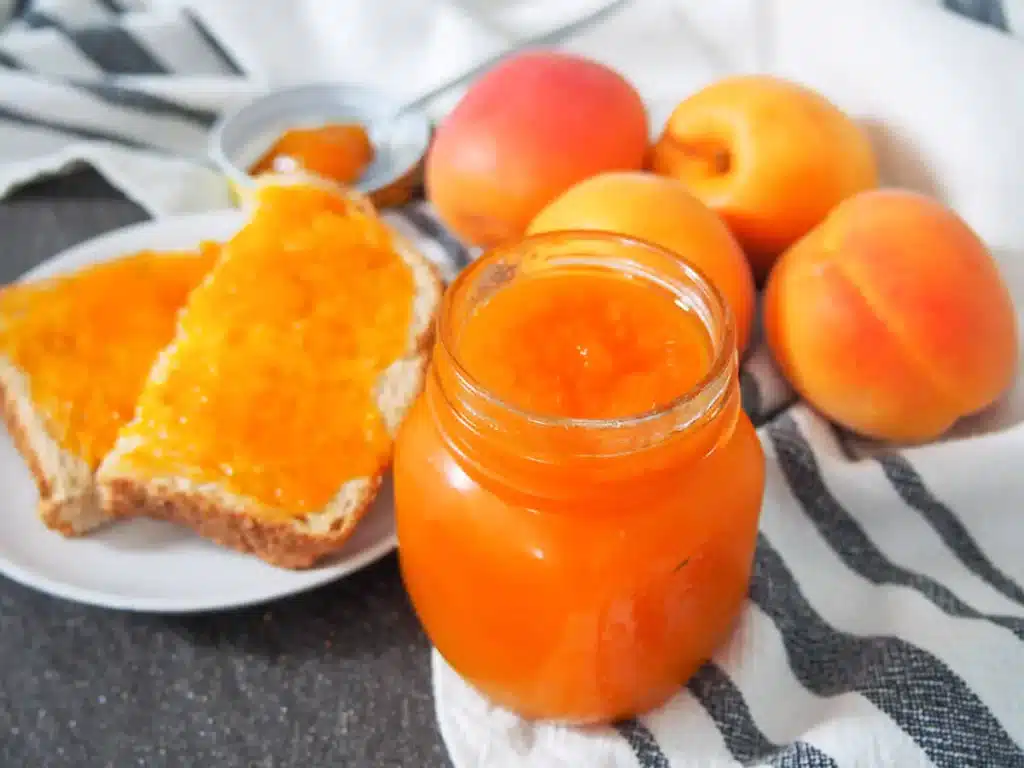 A view of a apricot jam in a glass jar with some jam on the toast and some apricots on the side