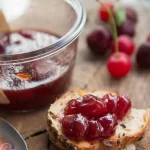 A view of cherry jam on a loaf of bread alongside a jam bowl