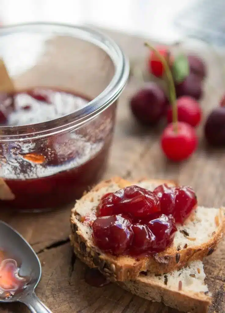 A view of cherry jam on a loaf of bread alongside a jam bowl