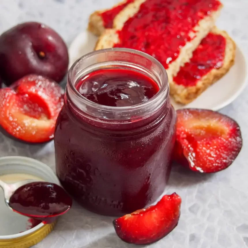 A view of plum jam inside a glass jar with plum cut on the side and jam on toast