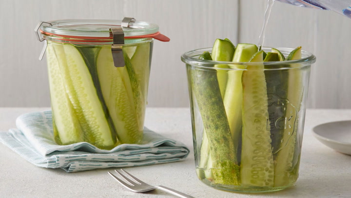 A view of two jars with brine solution being poured into one filled with cucumber pickle