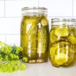 jars of dill pickle placed on a marble countertop along side dill seeds