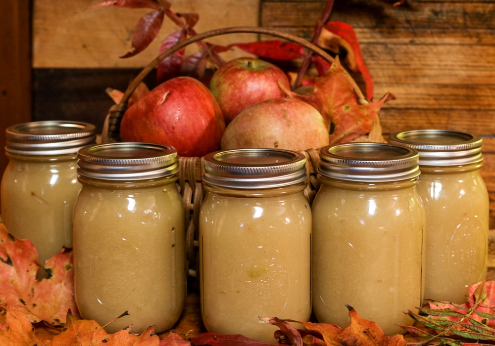 Glass Jars of homemade applesauce on a wooden board with autumn leaves and a basket of fresh apples