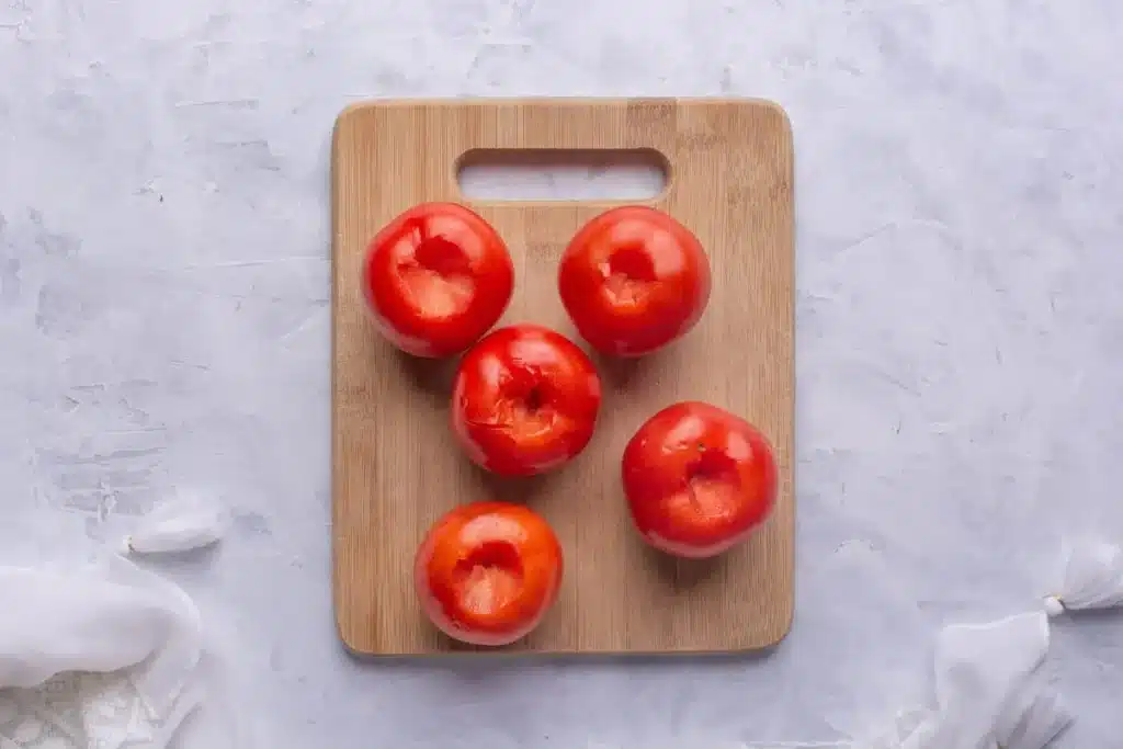 Five ripe tomatoes with their cores removed placed on a wooden cutting board ready for preparation or preservation