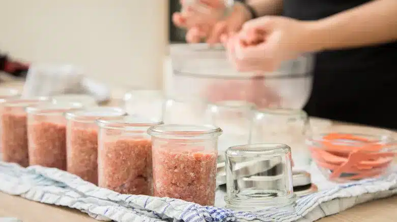 The image shows mason jars filled with ground meat with a person preparing more in the background The image shows mason jars filled with ground meat with a person preparing more in the background